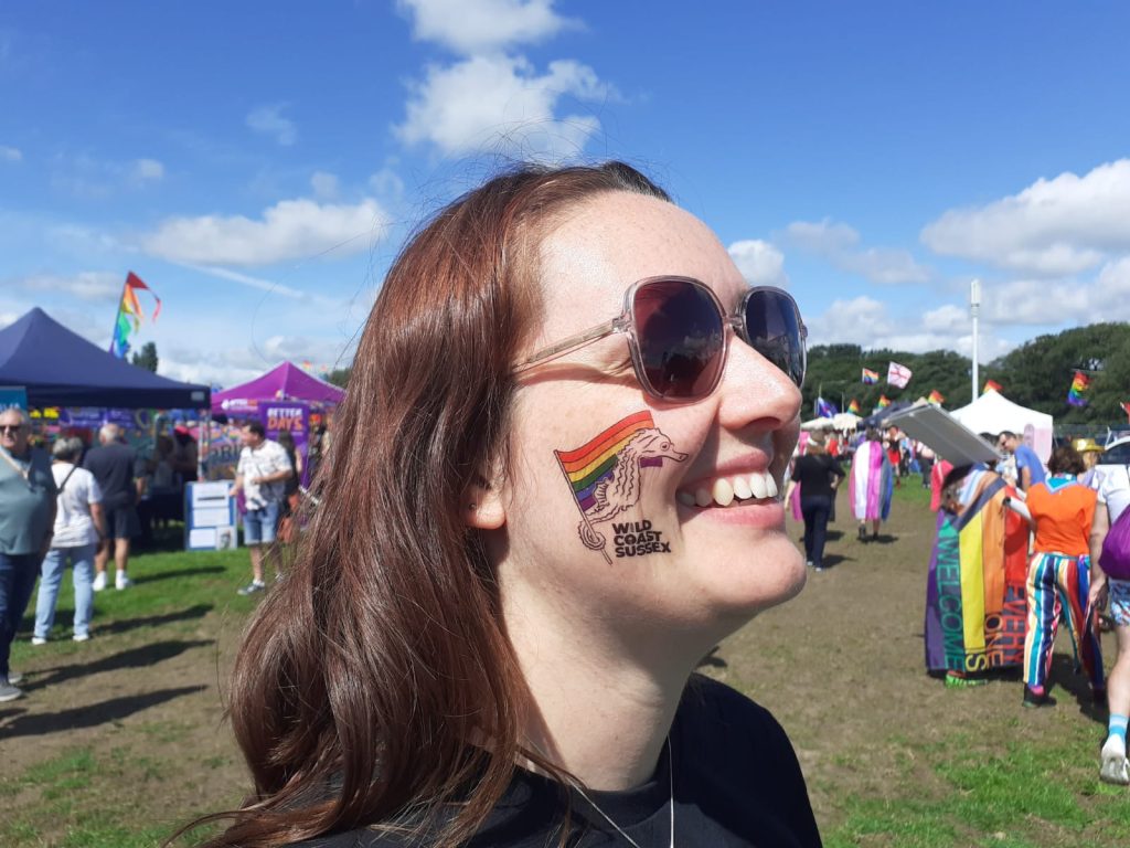A close up of a smiling womans face (sophie from the team) with a temporary tattoo of a seahorse holding a rainbow pride flag and the wild coast sussex logo