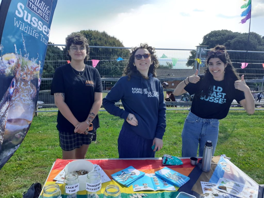 Three smiling people stand behind a table covered with the pride flag, go explore leaflets and marine species guides. One is a volunteer, two are team members wearing a wild coast sussex t shirt and a marine conservation society t shirt.