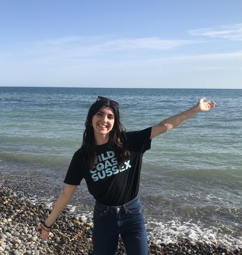 Sophie M is standing in front of the sea with her arms out and is smiling and wearing a Wild Coast Sussex T-shirt