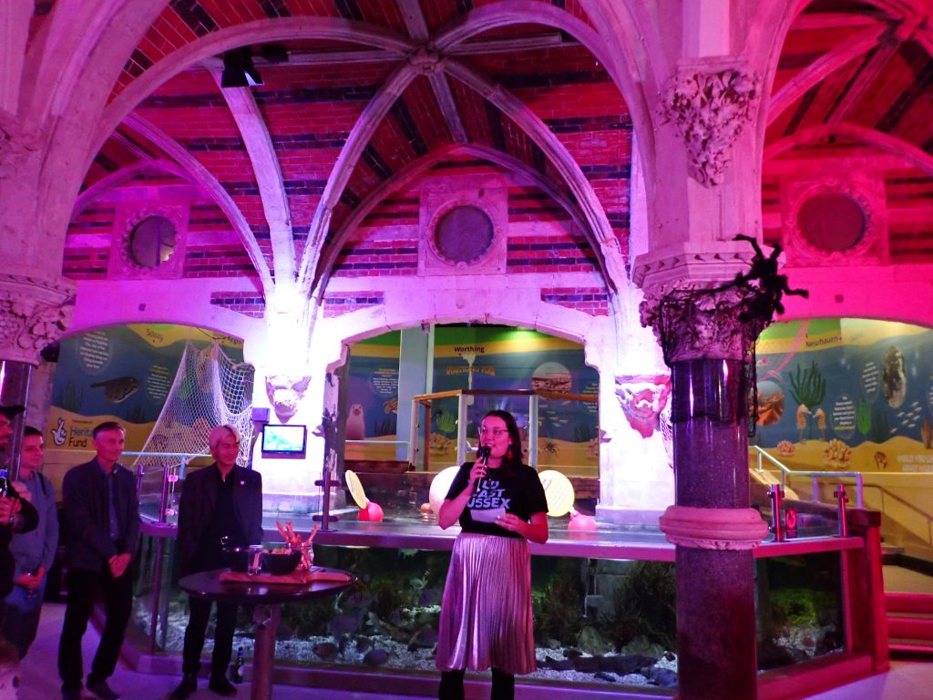 Sophie A is giving a talk in the Sealife centre, the lights are dimmed and red tinted and the ray tank in behind her.